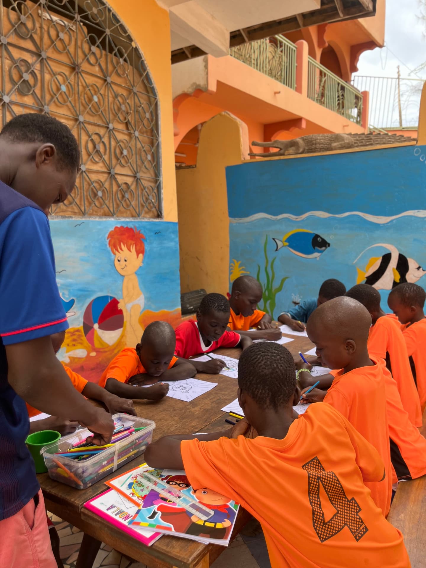Enfants en maillots orange dessinant autour d'une table devant une fresque marine