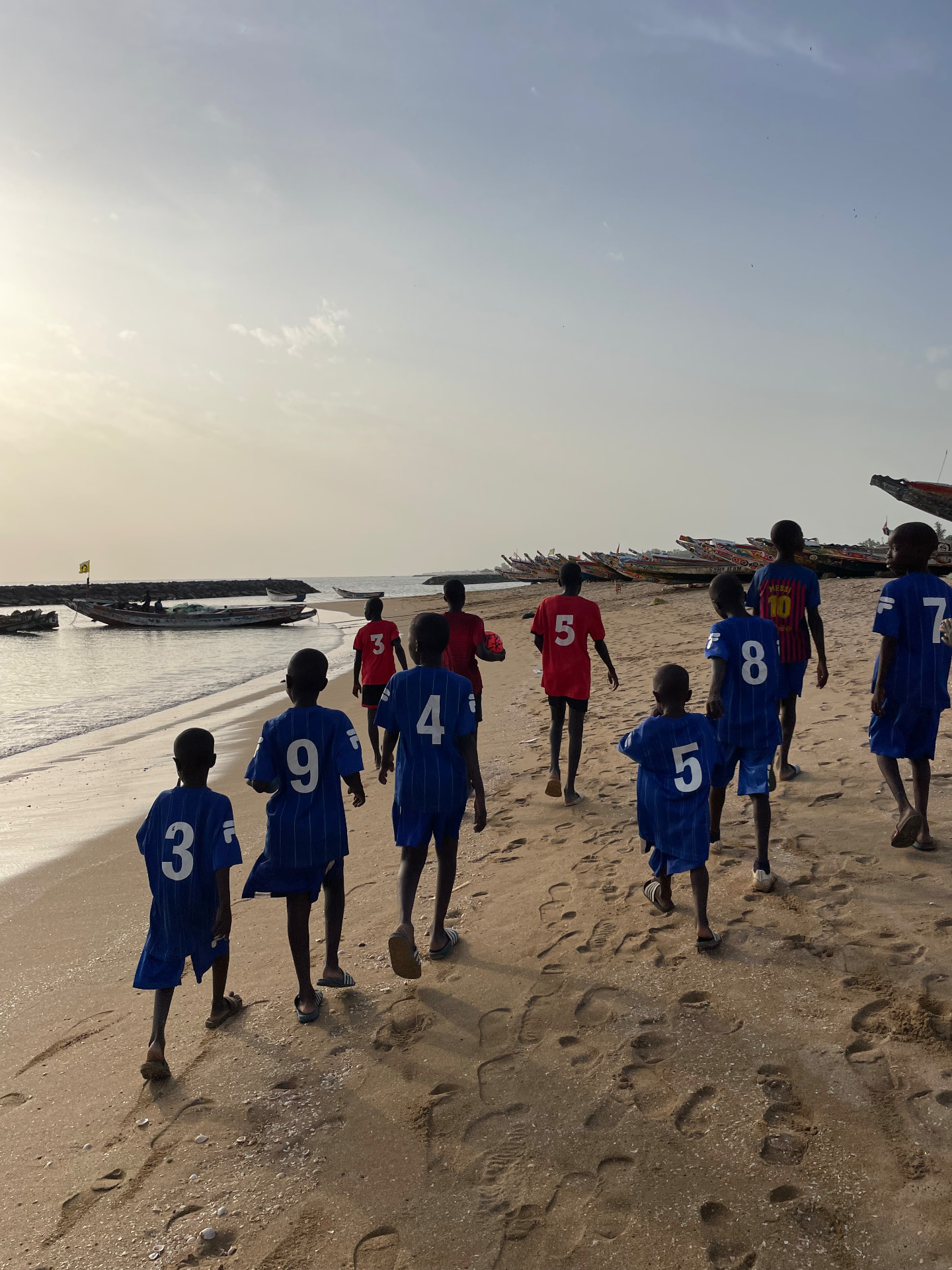 Enfants en maillots bleus et rouges marchant sur la plage de Mbour au coucher du soleil
